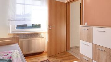 A bedroom with a wooden door, a radiator, a window with sheer curtains, a bed with a floral bedspread, and a wardrobe with multiple drawers.