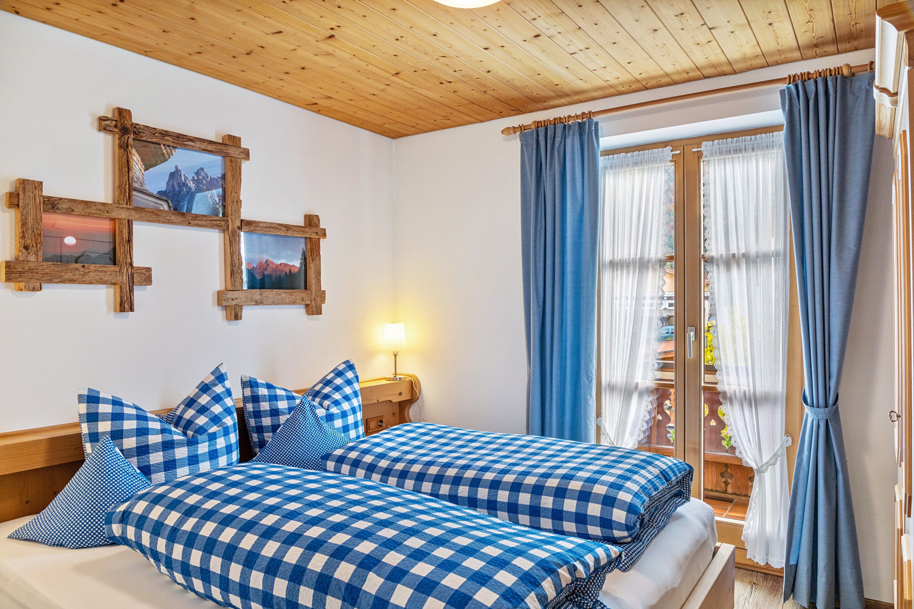 Ein Zimmer mit Holzbalkendecke, zwei Betten mit karierter Bettwäsche, einem Nachttisch mit Lampe und einem Fenster mit blauen Vorhängen.