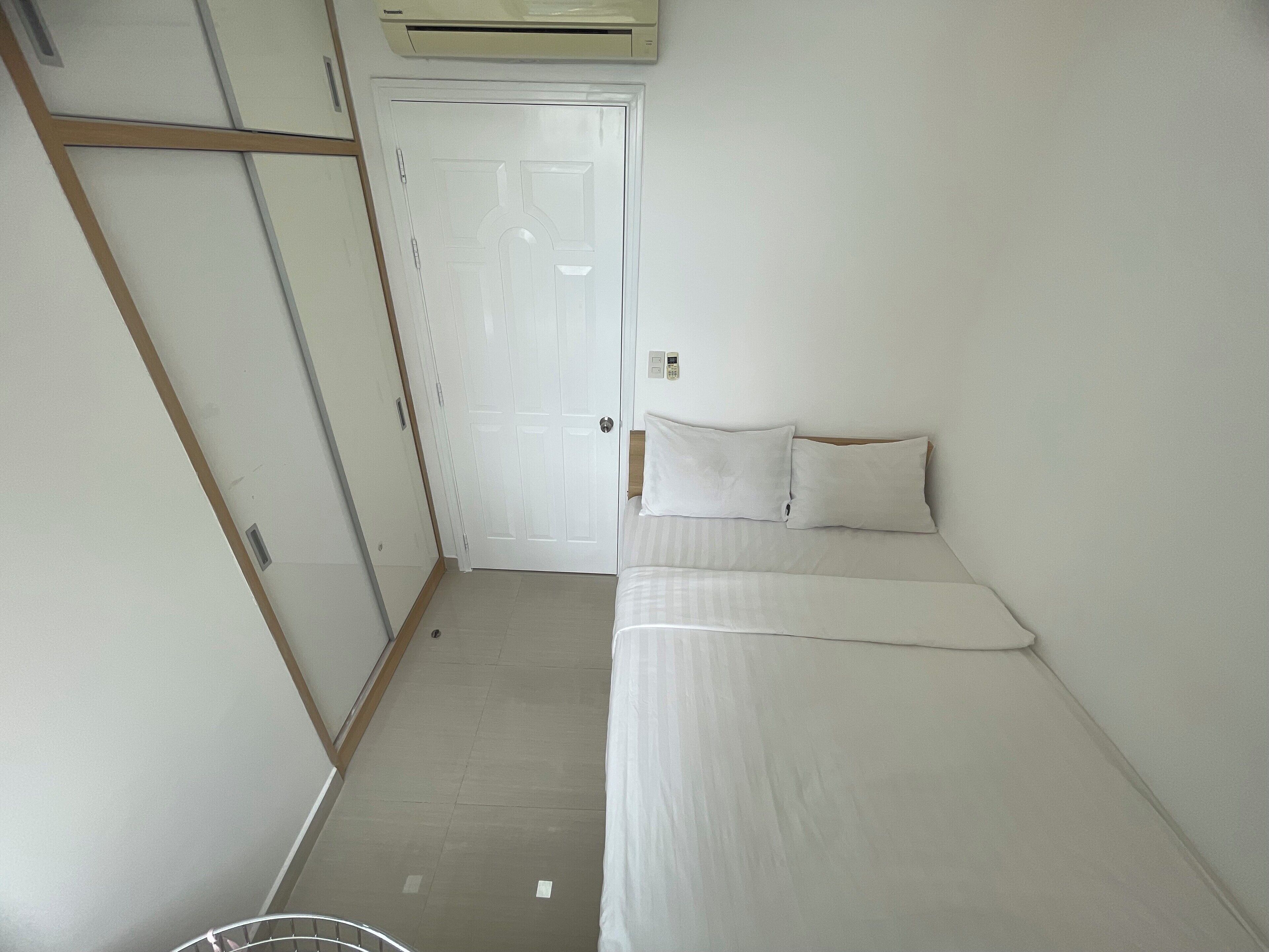 Une petite chambre blanche, avec un lit simple, une porte en bois et un mur blanc.