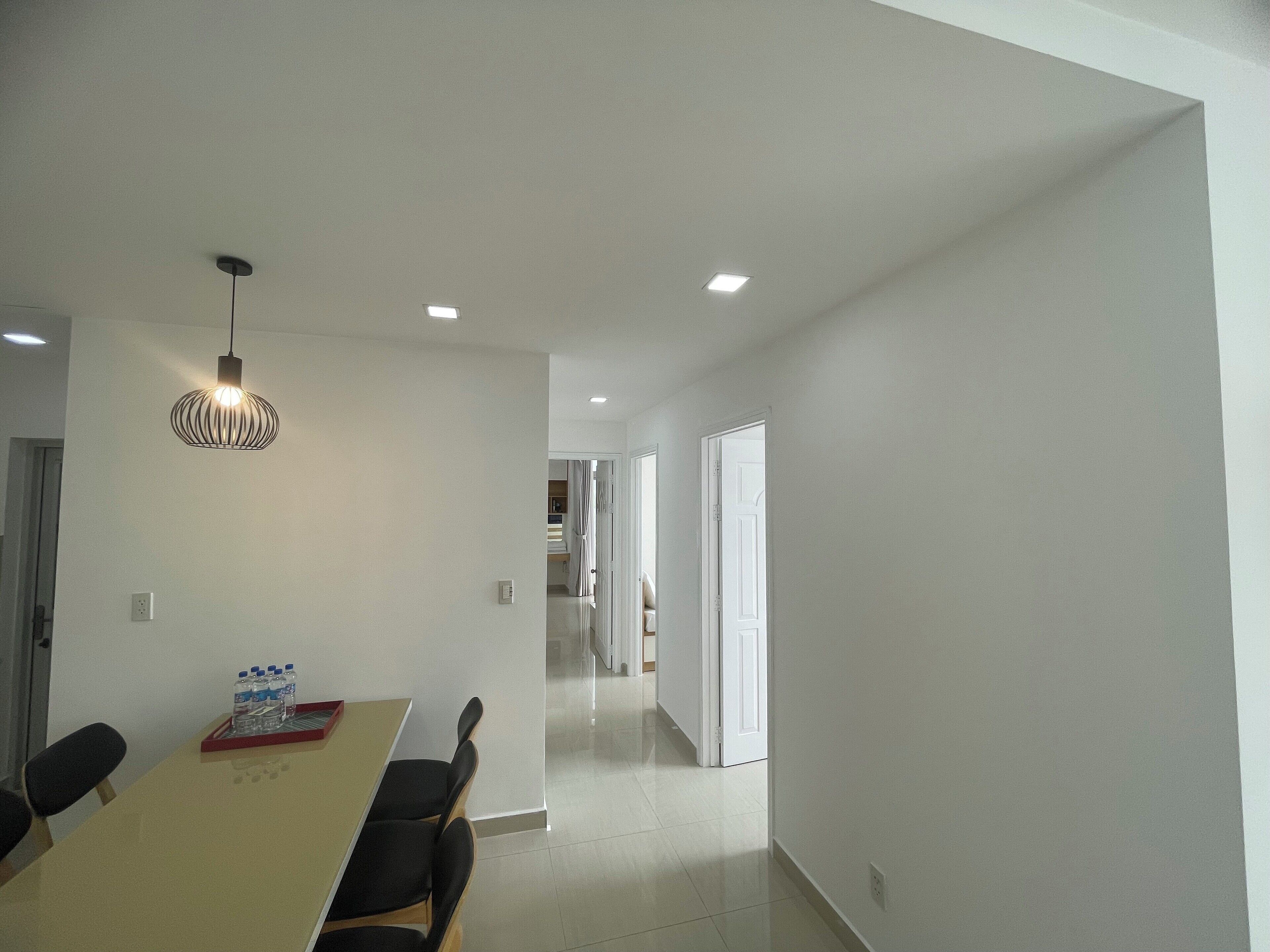 A modern dining area with a long table and chairs, a hanging light fixture, and a hallway leading to another room.