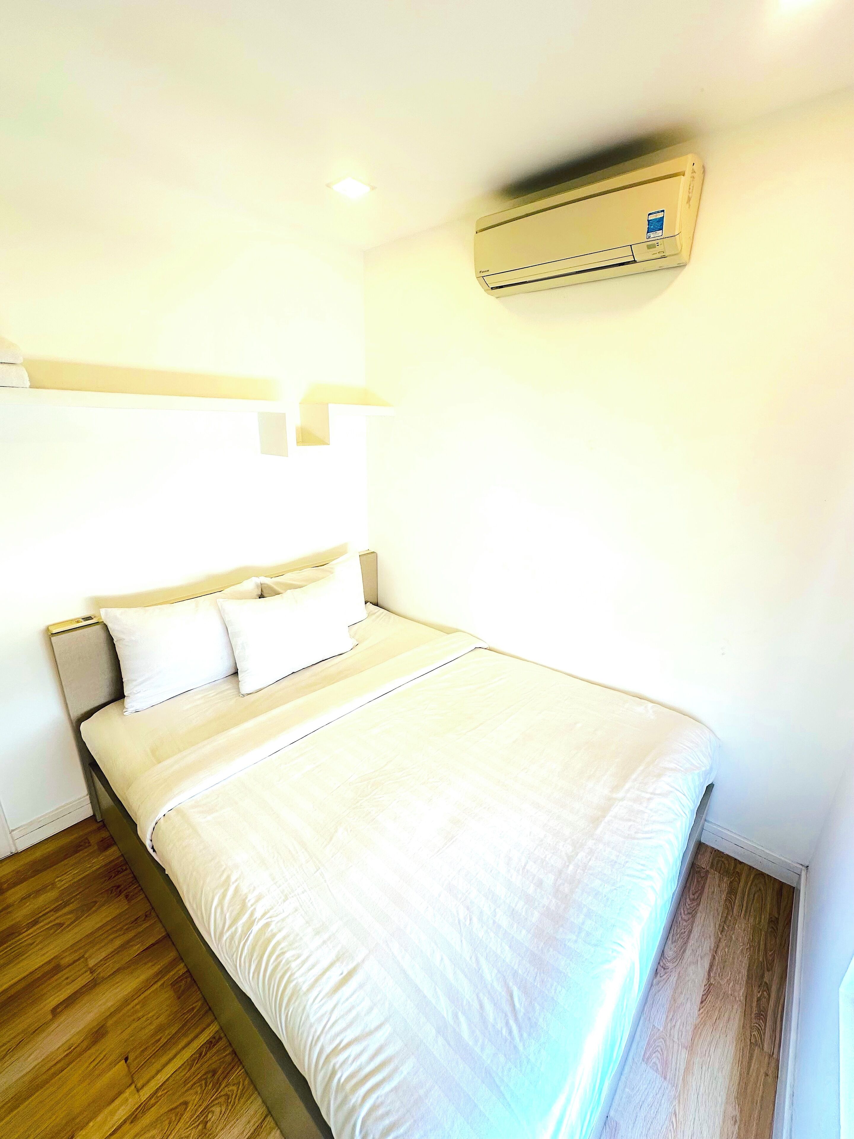 A neatly made bed with white bedding, a wooden floor, and a wall-mounted air conditioner.