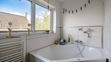 A modern bathroom with a large bathtub, a window with a view of a tiled roof, and a wall-mounted shower.