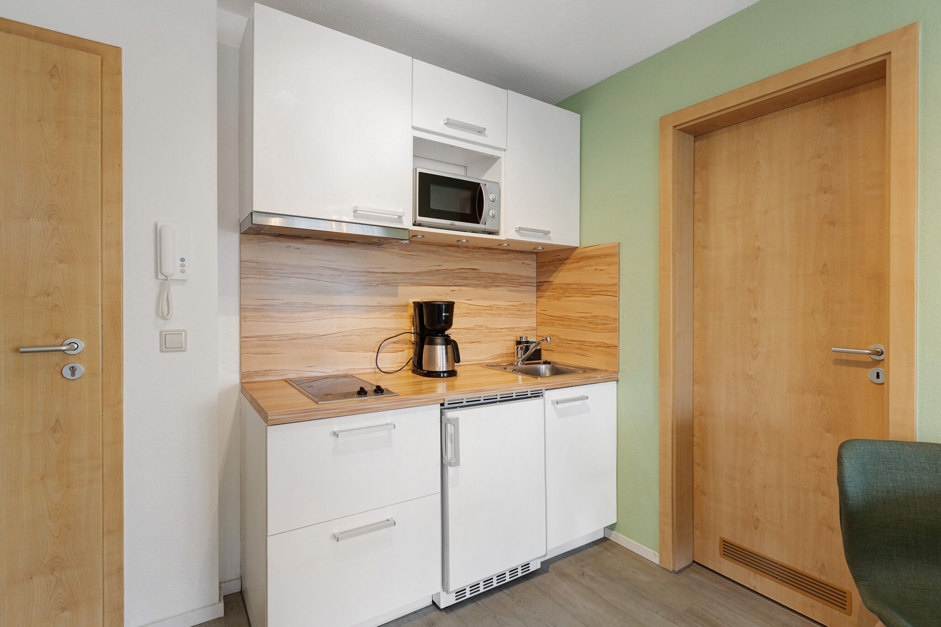 Una cocina compacta con armarios blancos, microondas, cafetera y lavamanos.