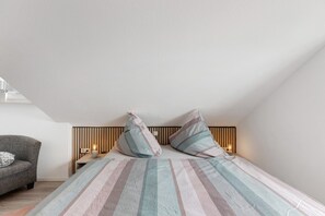 Ein Schlafzimmer mit einem Bett, zwei Kissen, einem Nachttisch, einer Lampe, einem Sessel und einer schrägen Decke.