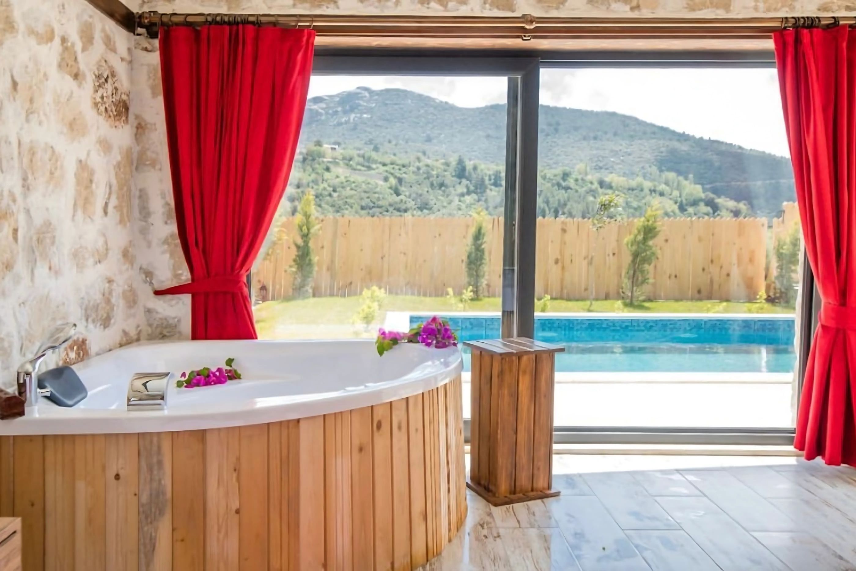 A bathroom with a wooden bathtub, a window with red curtains, and a view of a swimming pool and mountains.