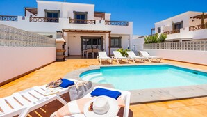 Un área de piscina con sillones de descanso y un edificio de fachada blanca y paredes con azulejos.