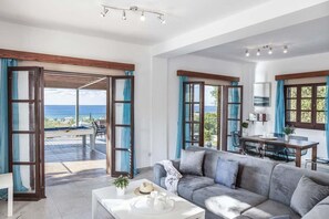 En rummelig opholdsstue med sofa, sofabord og spisebord. Store vinduer med blå gardiner giver udsigt til havet.