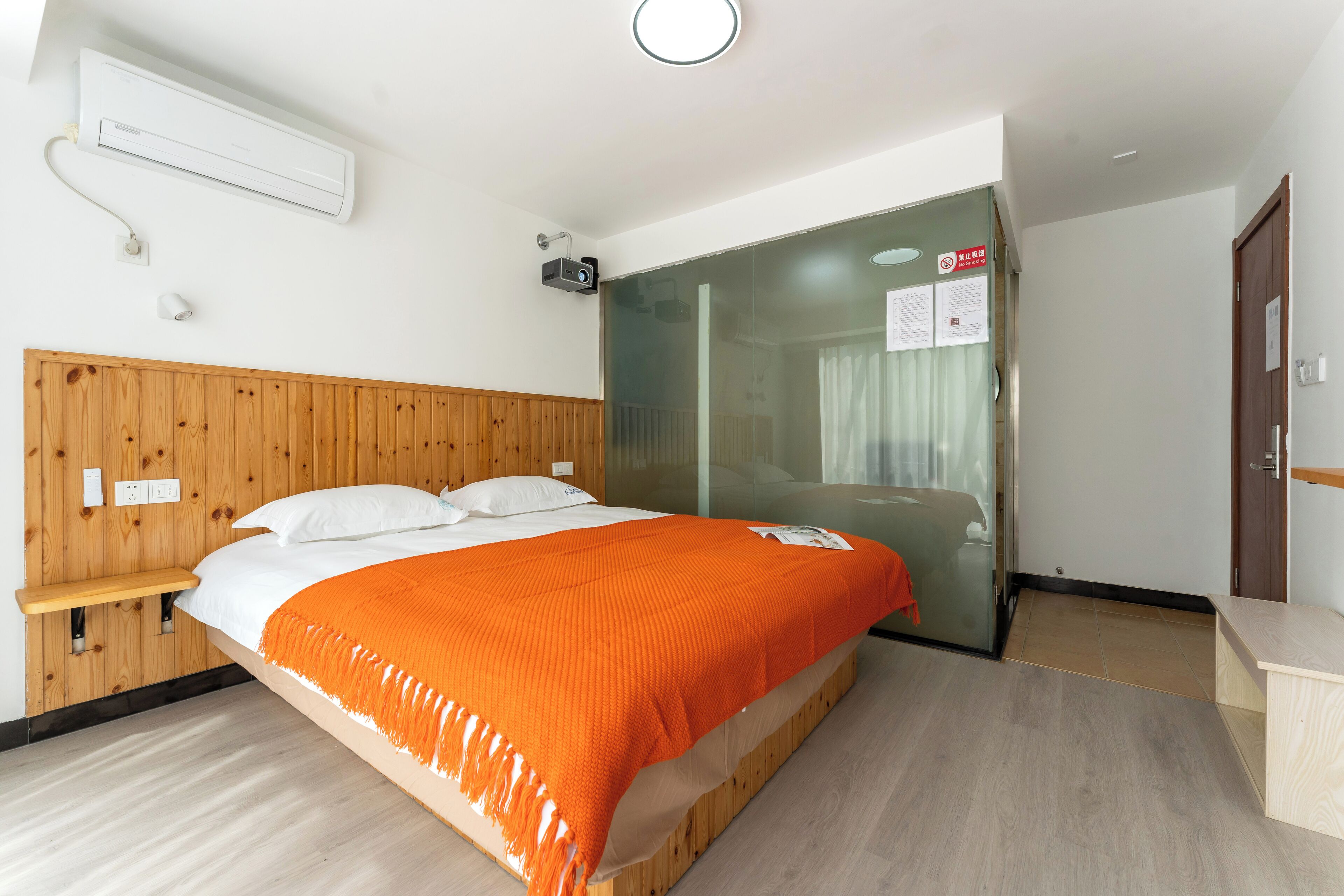 A hotel room with a wooden headboard, a bed with an orange blanket, a glass shower enclosure, and a wooden nightstand.
