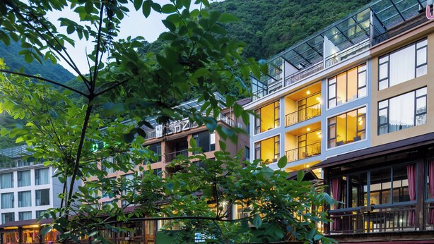 Ein mehrstöckiges Hotel mit Steinaußenfassade, vor dem ein Fluss fließt und dahinter Berge aufragen.