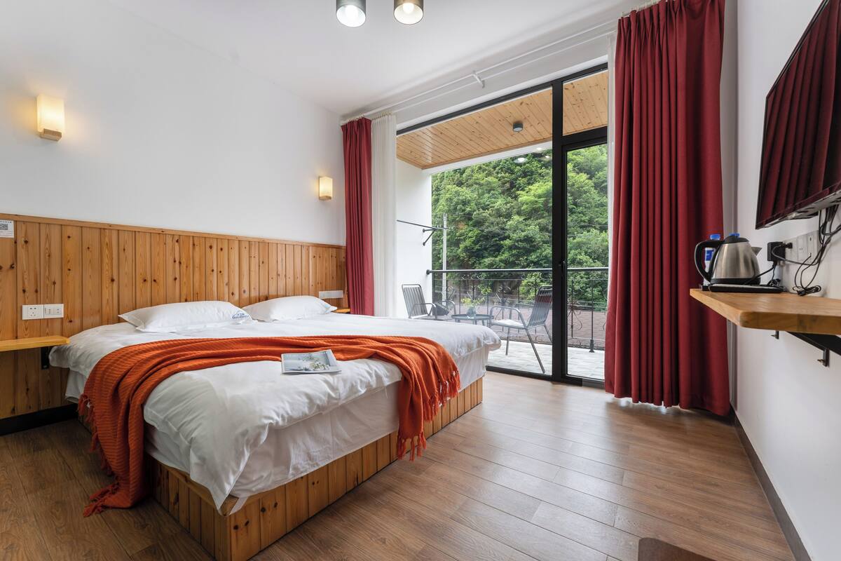 A modern bedroom with a wooden bed, white bedding, and a red blanket. There is a balcony with red curtains and a view of greenery.