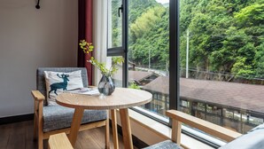 Ein modernes Zimmer mit einem großen Fenster, einem runden Holztisch, einem grauen Sofa mit einem Deer-Kissen und einer Vase mit Blumen.