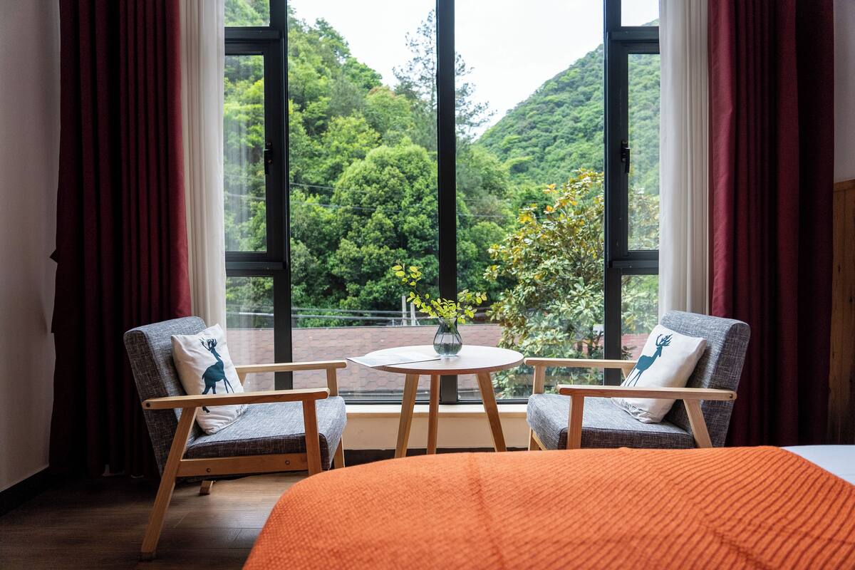 A room with a large window, two chairs, a small table, and a bed with an orange blanket.