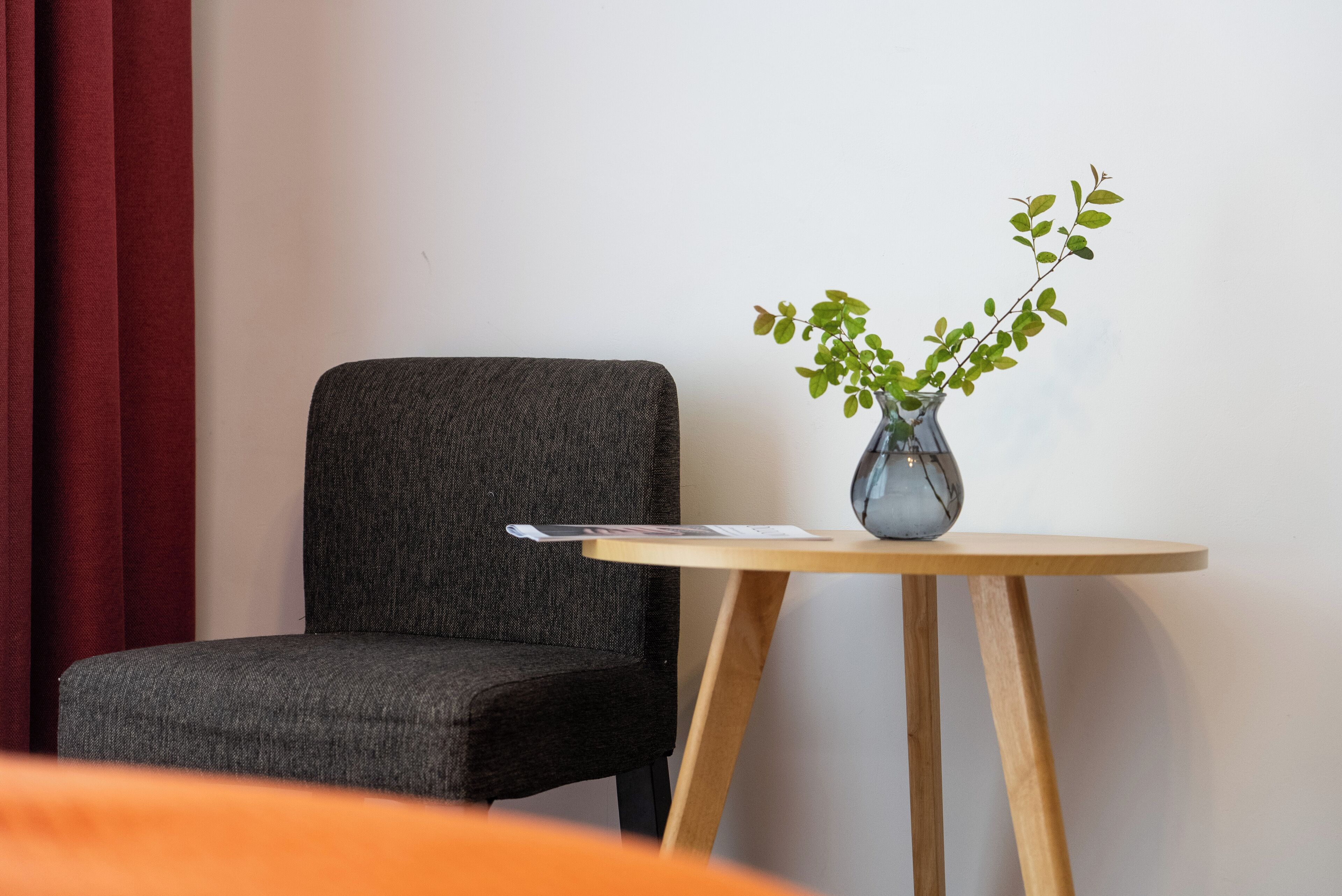Una habitación con una silla tapizada de gris, una pequeña mesa de madera con un florero de hojas verdes y una cortina roja.