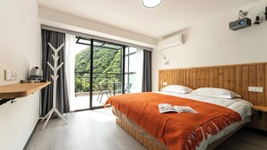 Ein modernes Hotelzimmer mit einem großen Bett, einem Holzkopfende, einem Nachttisch, einer Stehlampe und Blick auf üppiges Grün durch das Fenster.