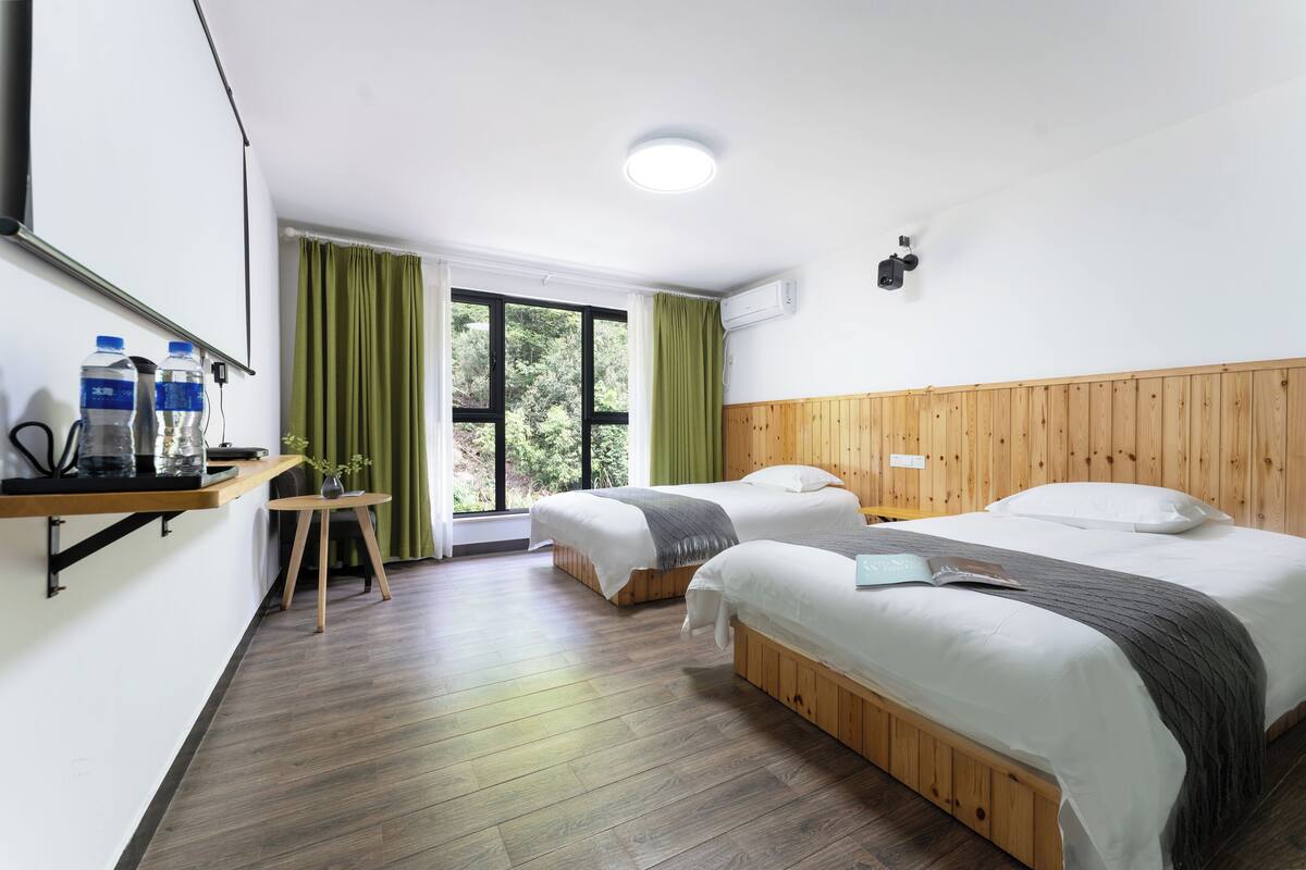 A hotel room with two beds, a wooden headboard, a small table with a vase, and a window with green curtains.