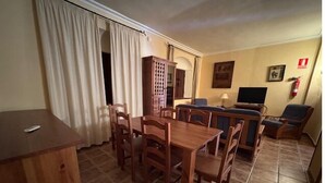 Een eetgedeelte met een houten tafel en stoelen, een klein kastje en een televisie op de achtergrond.