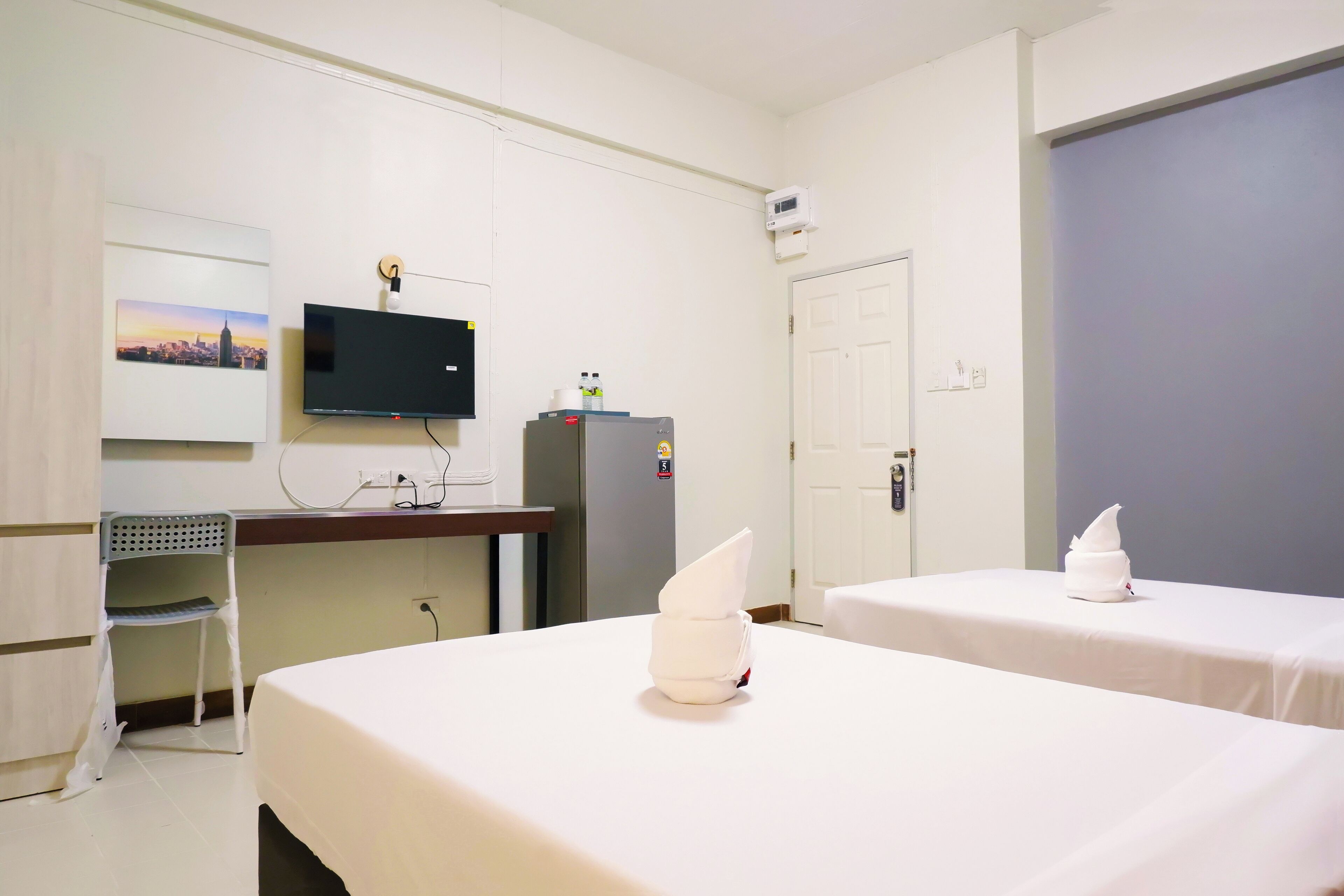 A hotel room with two beds, a desk with a chair, a television, a refrigerator, and a door.