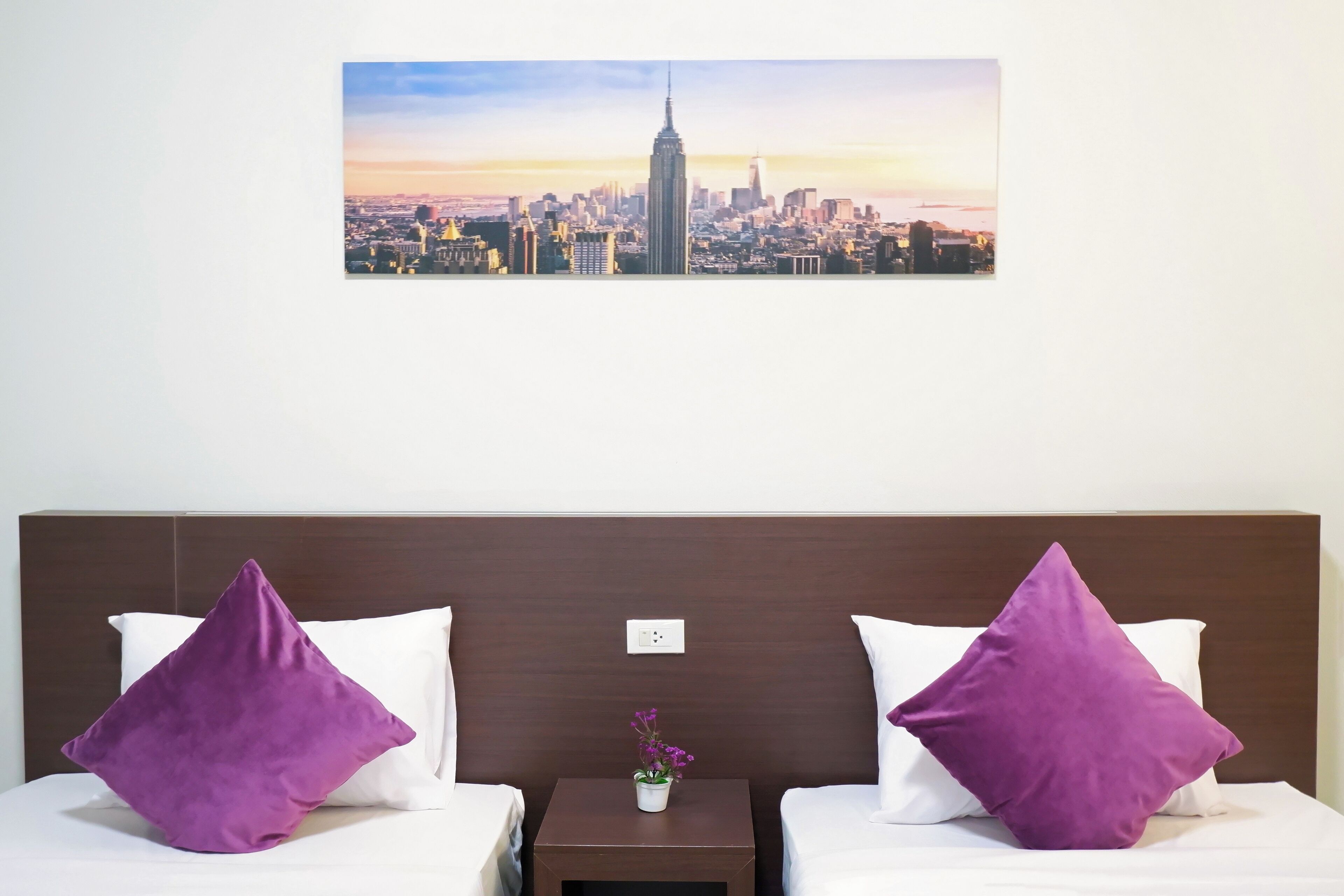 Ein Hotelzimmer mit zwei Betten, violetten Kissen, einem kleinen Tisch mit Blumenvase und einem Stadtblick an der Wand.