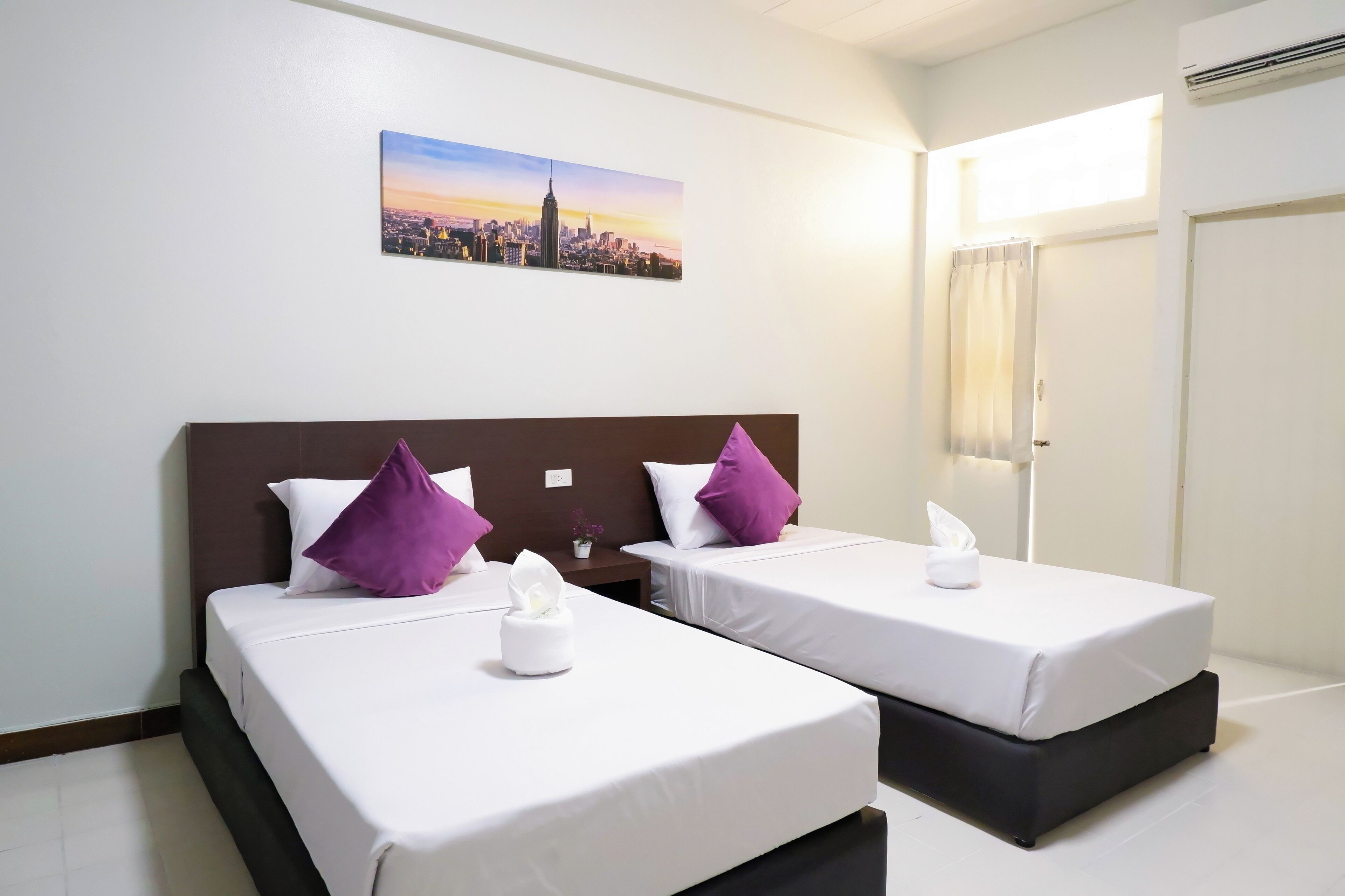 Une chambre d’hôtel avec deux lits, du linge de lit blanc, des oreillers violets, un tableau représentant un paysage urbain, une fenêtre avec des rideaux et un appareil de climatisation.