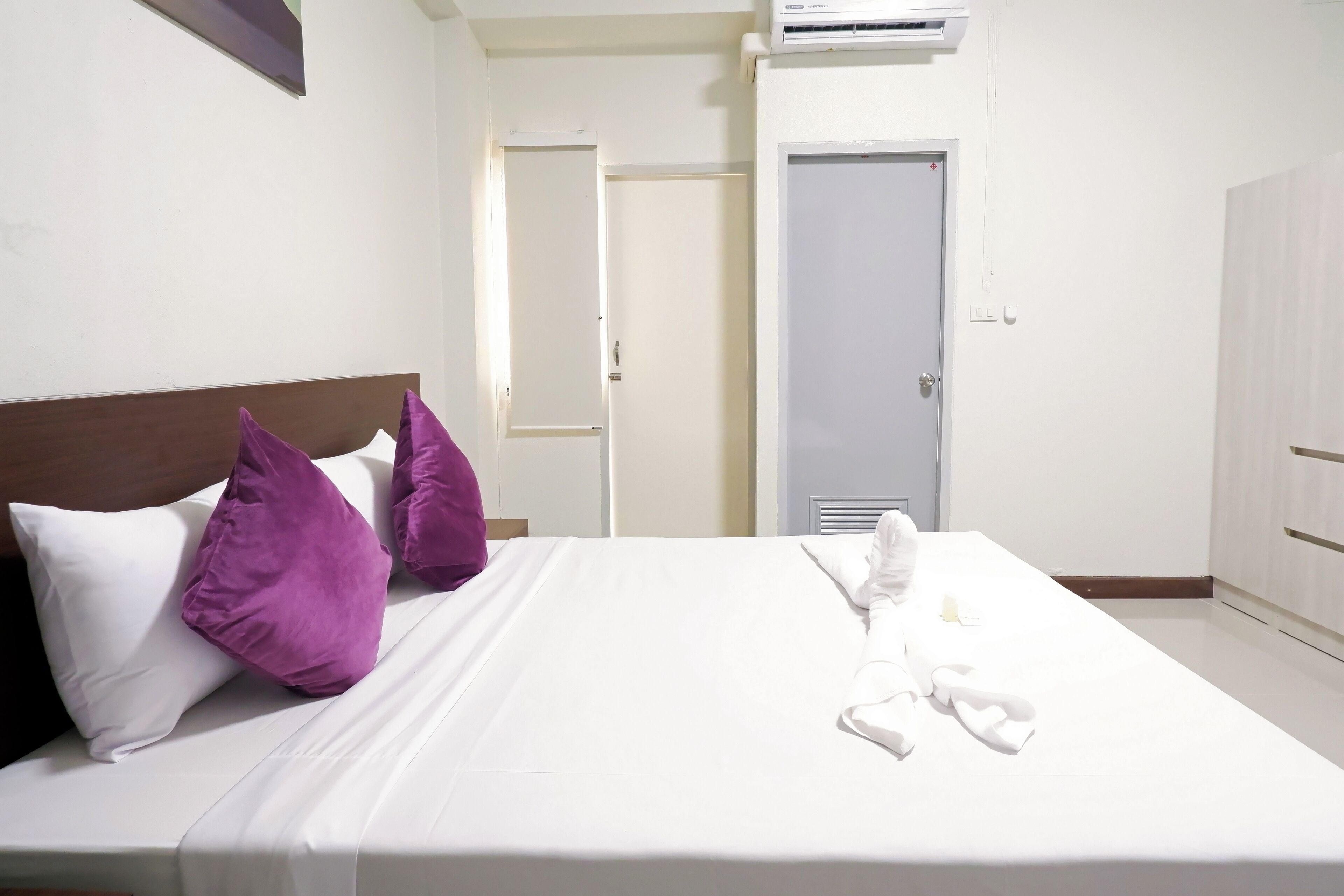 Une chambre d’hôtel avec un lit, du linge de lit blanc, des oreillers violets, une serviette blanche et une armoire blanche.