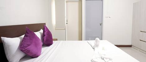 Habitación de hotel con cama, ropa de cama blanca, almohadas moradas, toalla blanca y armario blanco.