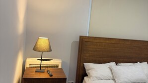 Een slaapkamer met een nachtkastje van hout, een lamp, een afstandsbediening en een bed met wit beddengoed.