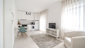 Un salon moderne comprenant un canapé, un téléviseur posé sur un meuble, un coin repas avec une table et des chaises, et un coin cuisine équipé d’appareils électroménagers et d’un lave-linge.