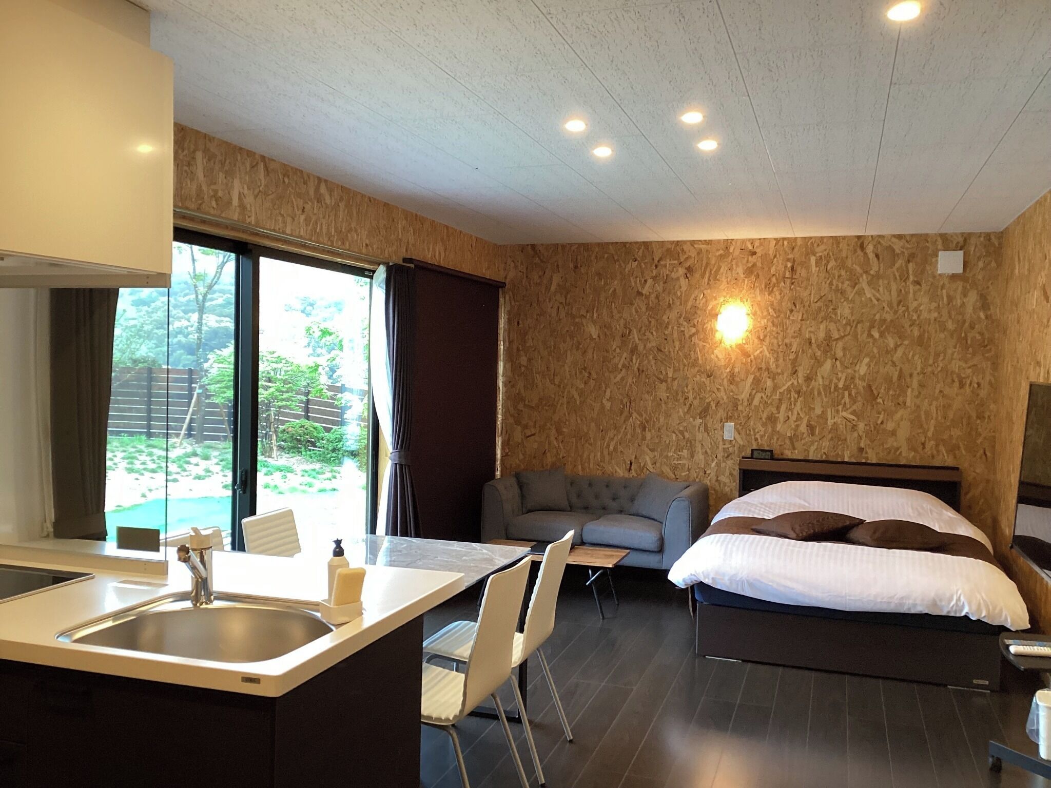A modern hotel room with a bed, a sofa, a dining area with a sink and chairs, and a large window with a view of greenery.