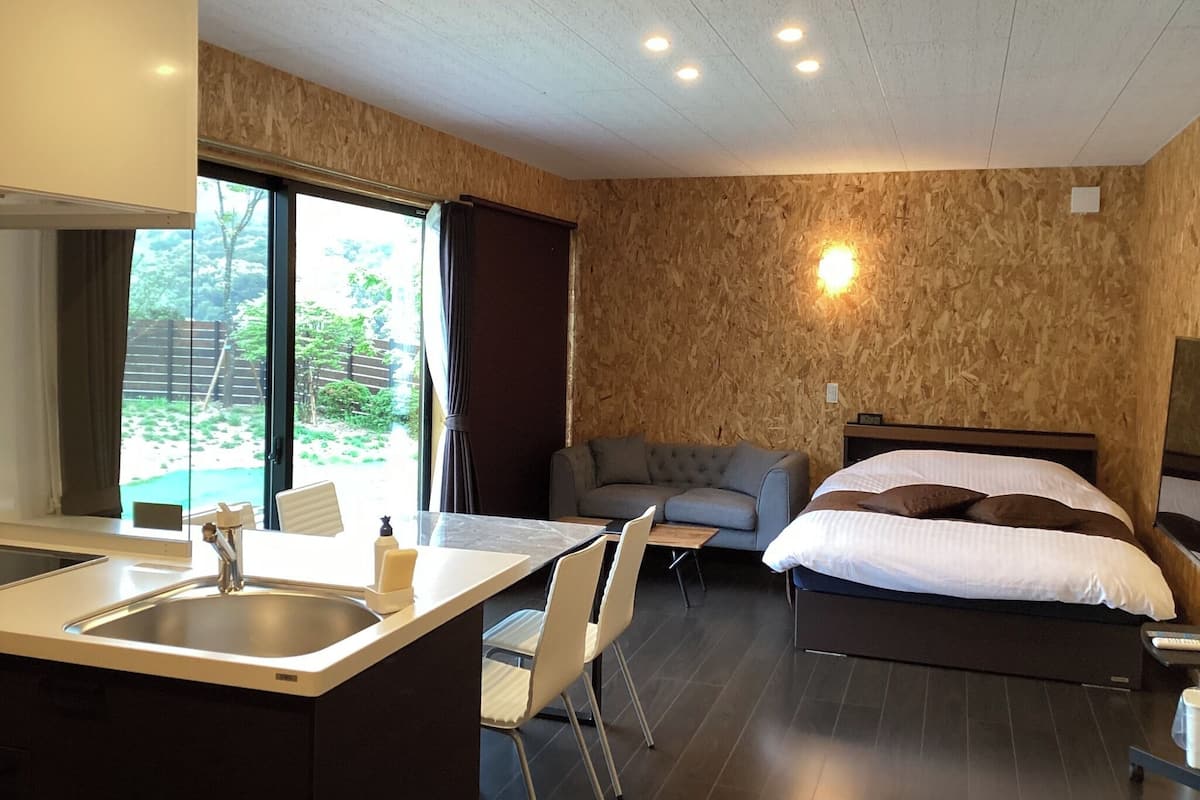 Una camera d'albergo moderna con un letto, un divano, un'area pranzo con un lavello e sedie, e un'ampia finestra con vista sul verde.