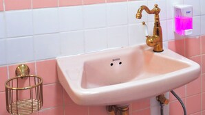 A bathroom with a pink tiled wall, a white sink with a gold faucet, and a round mirror above the sink. - Sweety Secret Guesthouse (Bangkok)
