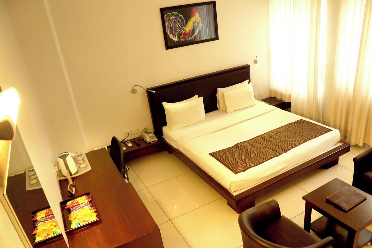 A hotel room with a bed, bedside table, chair, desk, and a painting on the wall.