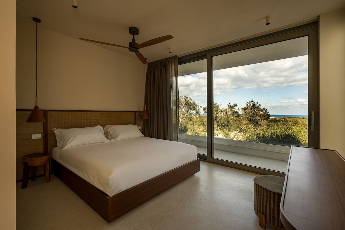 A bedroom with a large bed, a ceiling fan, a view of trees and sky through a sliding door, and a small table.