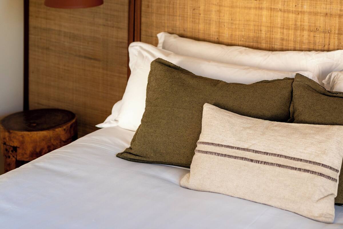 A neatly made bed with a wooden headboard, a hanging red pendant light, and a small wooden stool beside the bed.