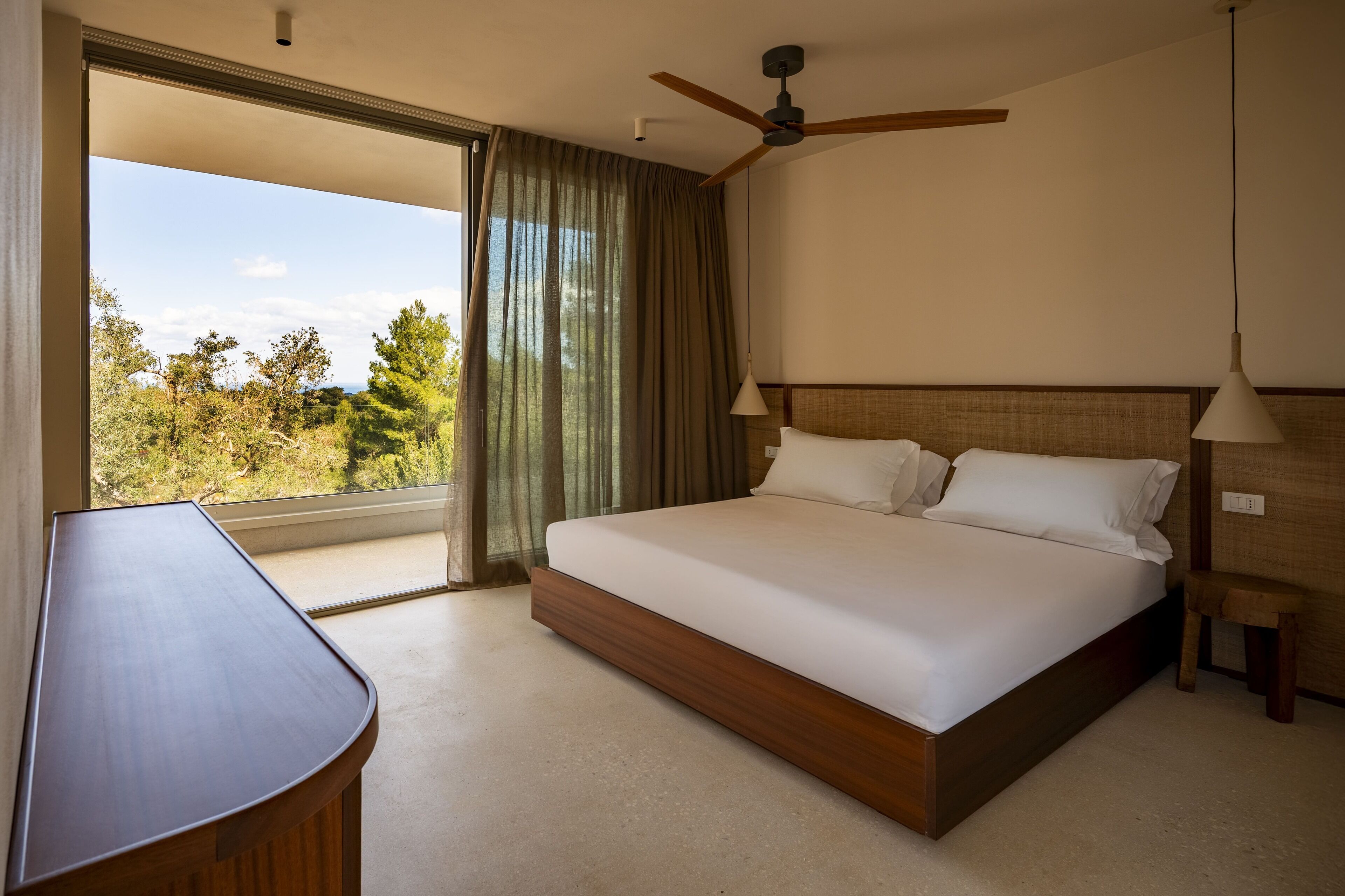 Un dormitorio con una cama grande, un ventilador de techo, un escritorio y vistas a árboles por la ventana.