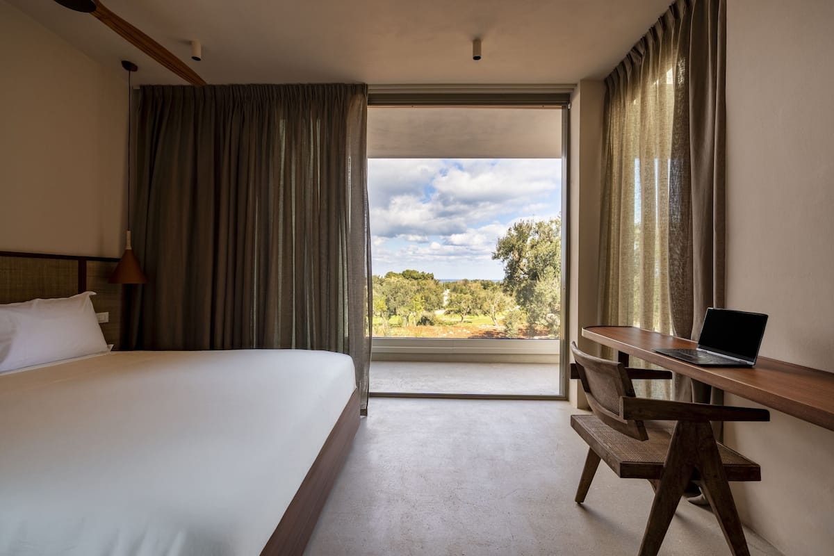 A hotel room with a bed, a desk with a laptop, and a view of the outdoors.