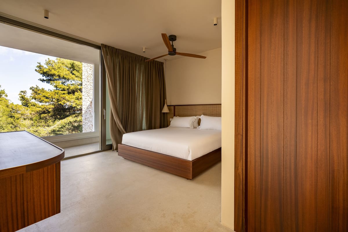 A modern hotel room with a large bed, a ceiling fan, and a view of trees through a large window.