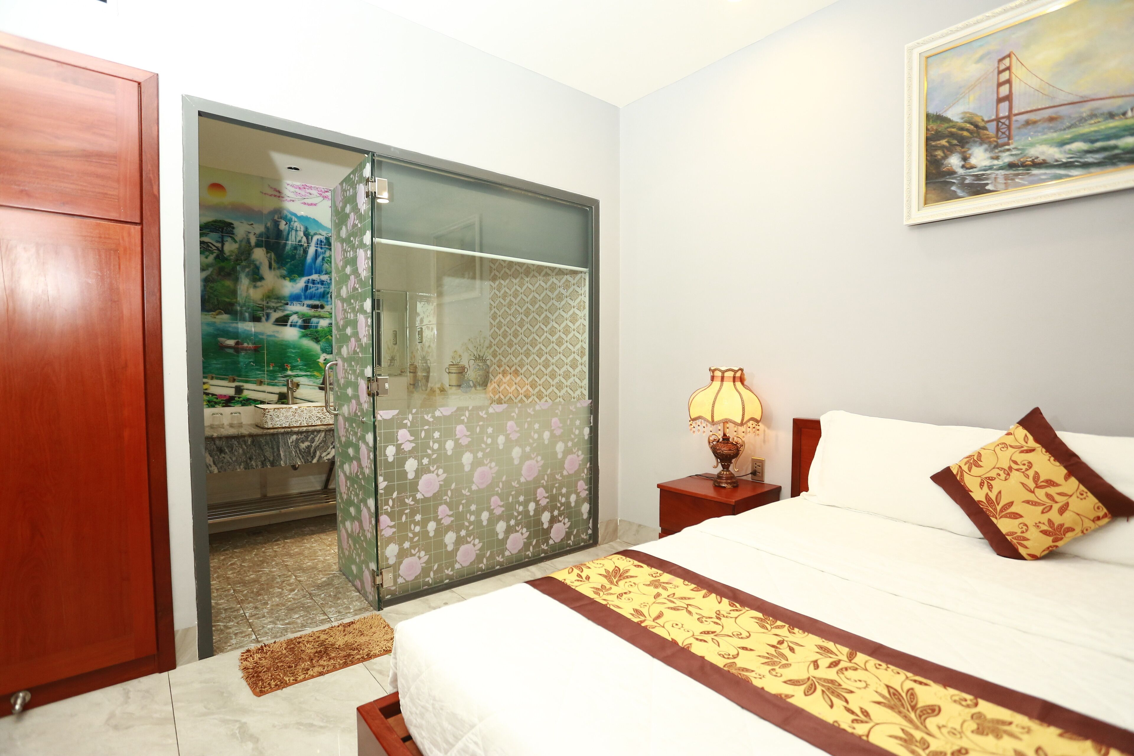 A hotel room with a bed, a nightstand, a lamp, a painting of a bridge, and a bathroom with a shower and sink.