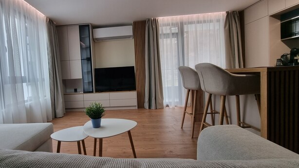 A modern living room with a flat-screen TV, two bar stools, a small table with a plant, and a sofa.