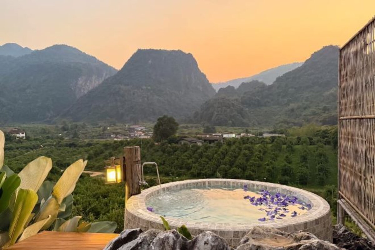 A hot tub with a mountain view, surrounded by greenery and a wooden deck.