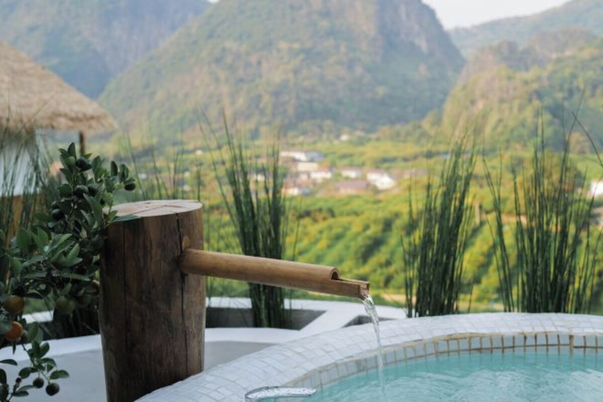 A pool with a bamboo water spout, surrounded by greenery and a mountainous landscape in the background.