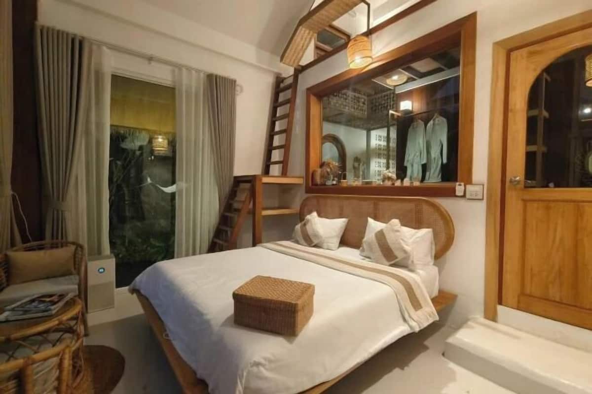 A bedroom with a bed, a wooden headboard, a mirror, a window with curtains, and a wooden door.