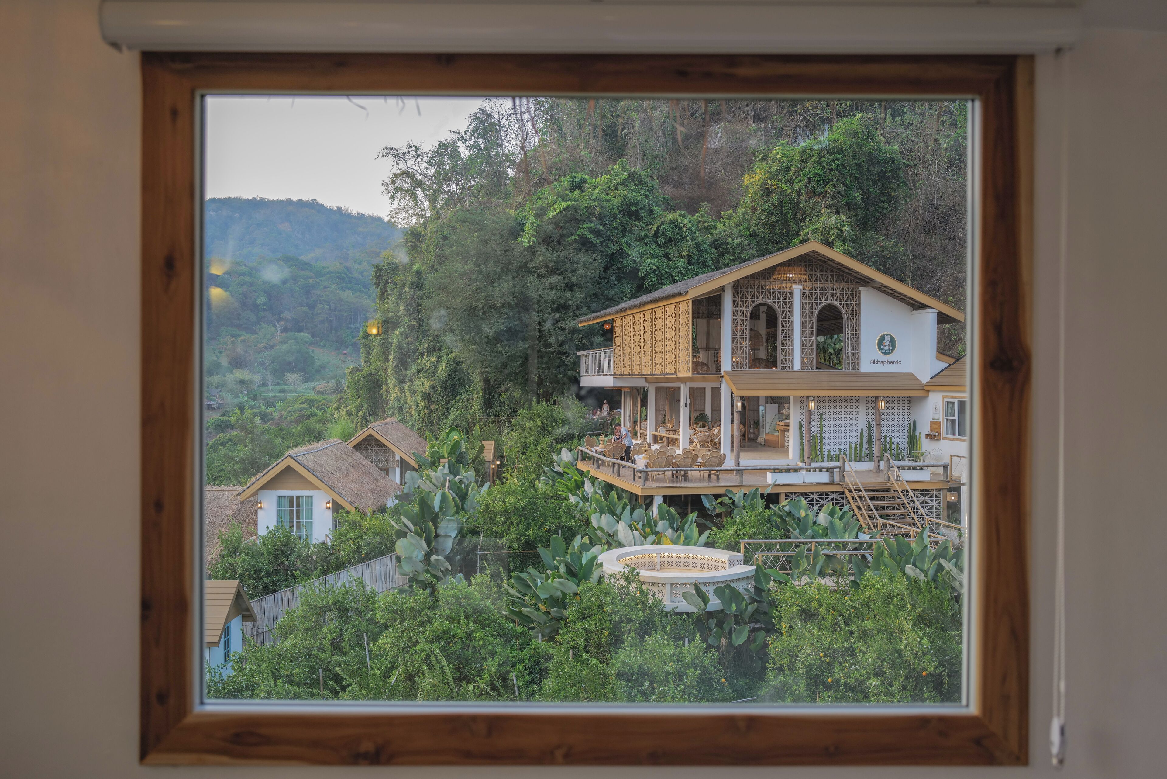 Una casa de dos pisos con balcón, rodeada de exuberante vegetación y colinas.