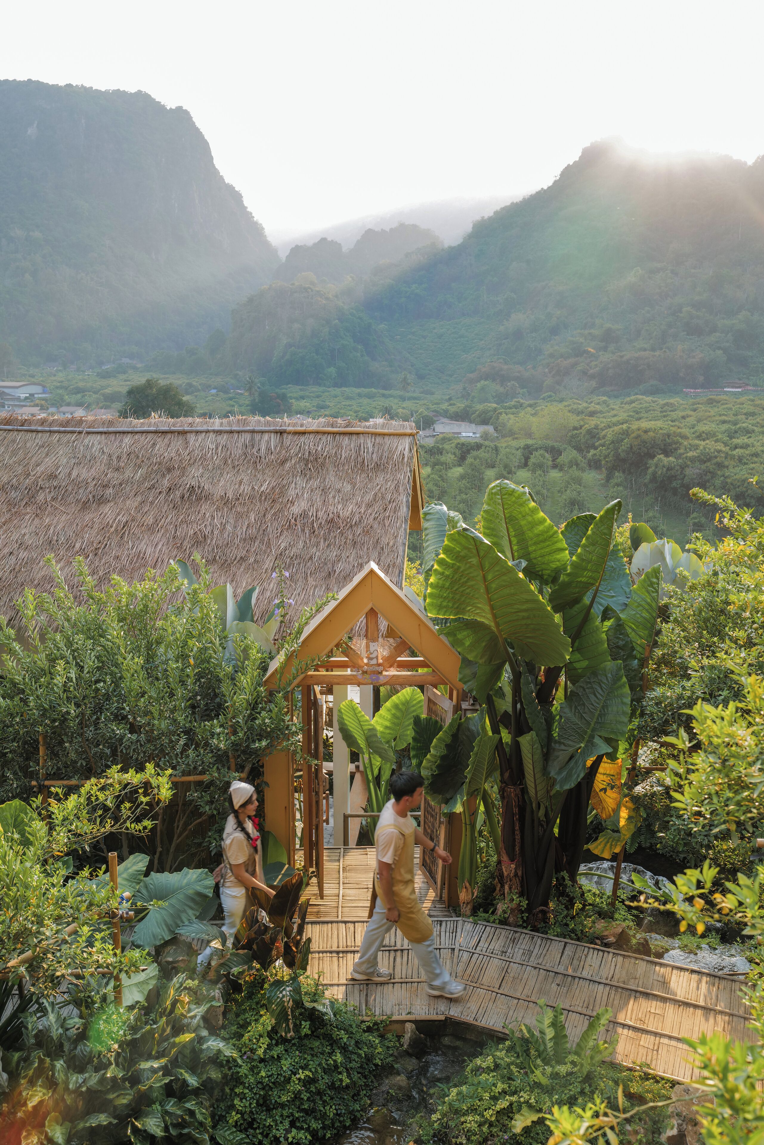 A rustic wooden bridge leading to a thatched-roof structure surrounded by lush greenery and mountains in the background.