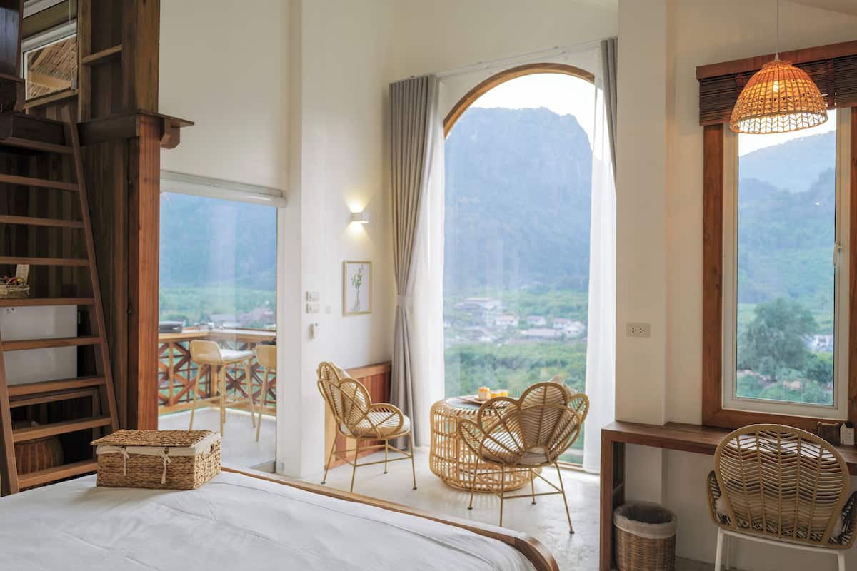A bedroom with a large bed, a wooden ladder, wicker furniture, and a view of a mountainous landscape through a large window.