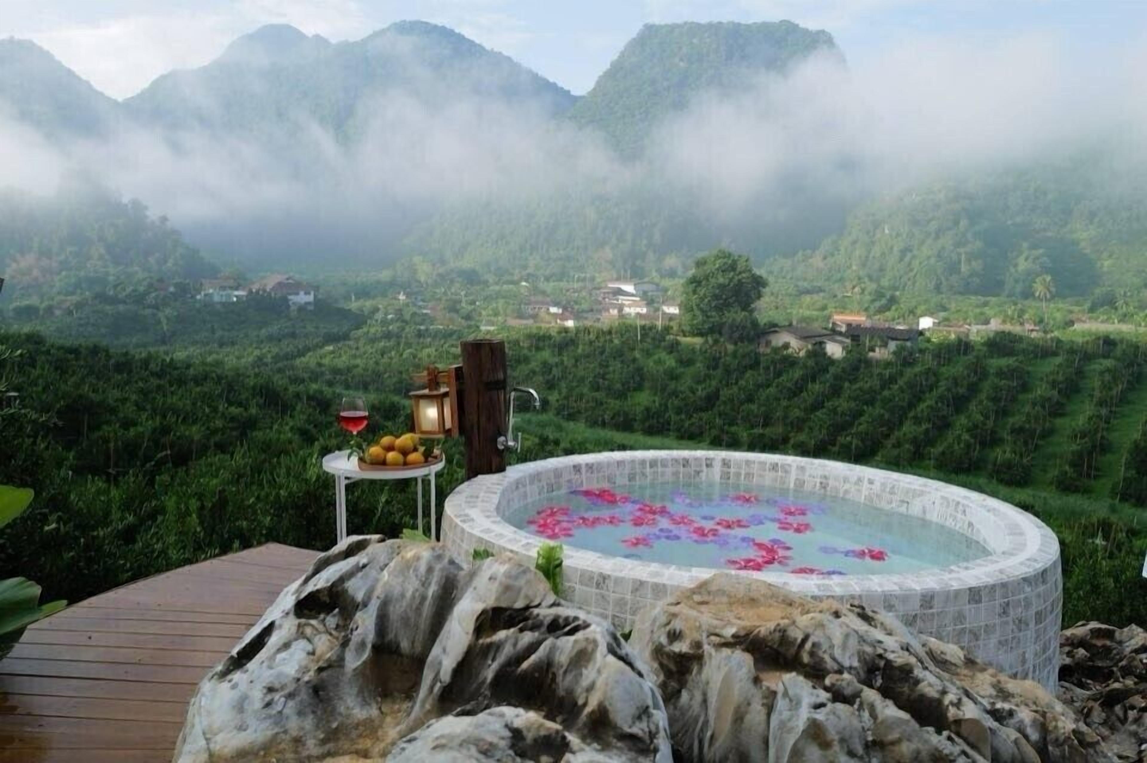 A hot tub surrounded by a wooden deck, with a table of fruits and a bottle of wine nearby.