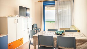 A dining area with a table set for four, a television on a cabinet, and a view of the sea through a window with curtains.