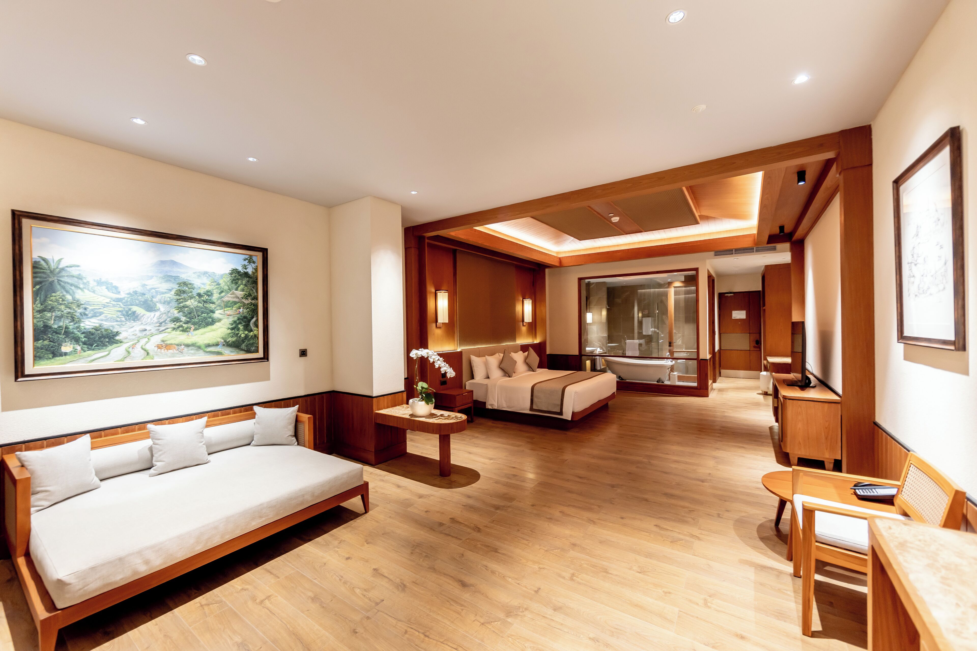 A spacious hotel room with a wooden floor, a large bed, a sofa, a small table with a vase, and a painting of a landscape.