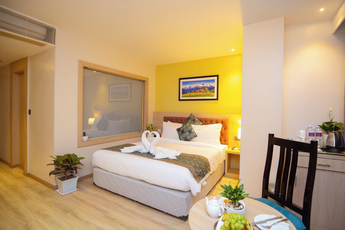 A hotel room with a large bed, a bedside table with a lamp, a small table with fruits and a teapot, and a painting on the wall.