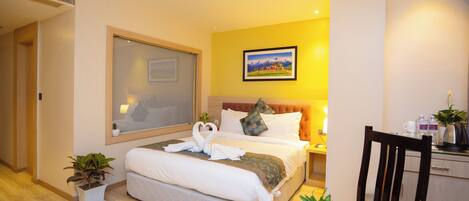 Una habitación de hotel con una cama grande, una mesita de noche con una lámpara, una pequeña mesa con frutas y una tetera, y un cuadro en la pared.