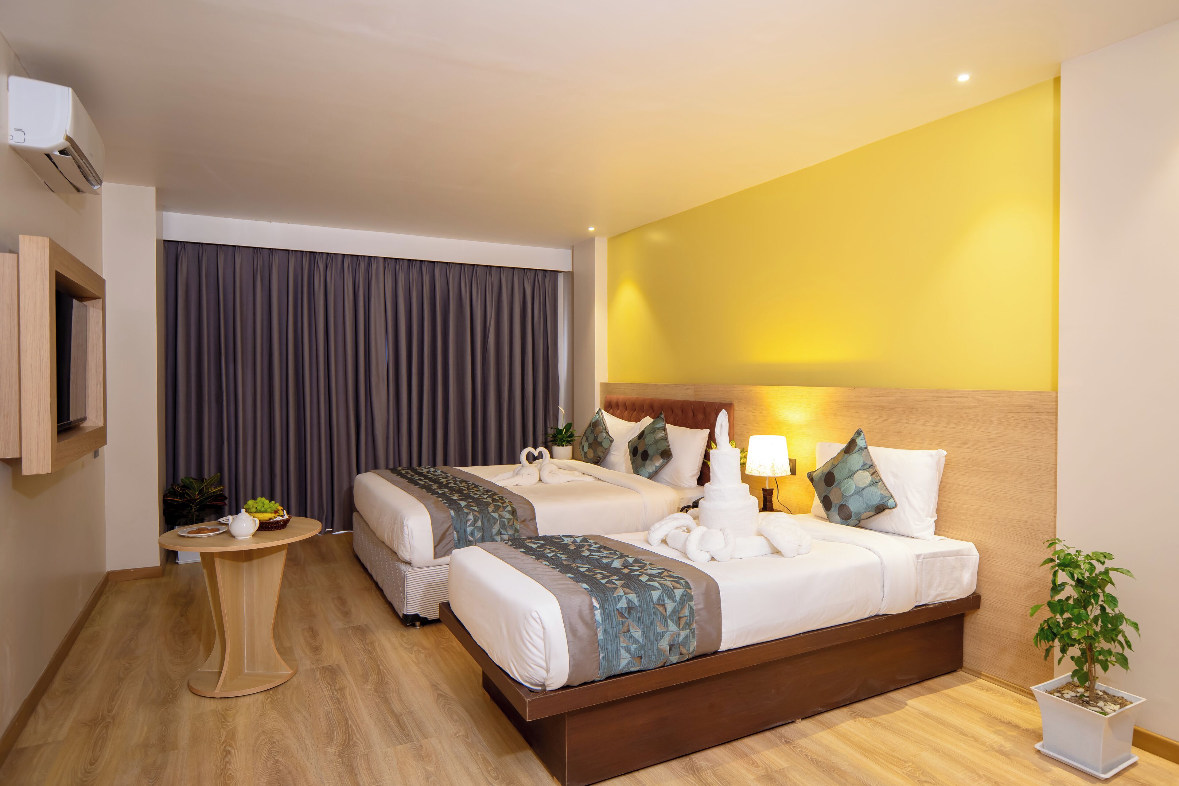 A hotel room with two beds, a small table, a potted plant, and a TV mounted on the wall.